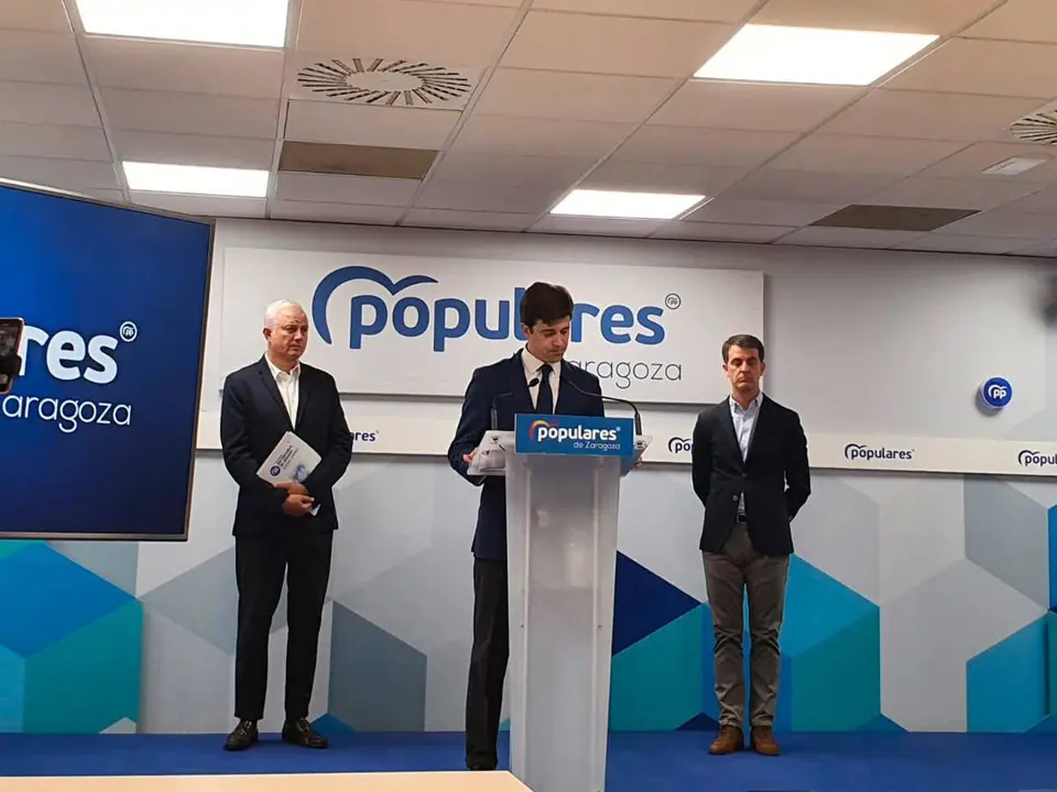 El presidente del PP en Zaragoza Ram&oacute;n Celma en el atril del partido. Detr&aacute;s, los dos diputados populares por la provincia de Zaragoza. Fondo blanco y azul con un cartel del partido.