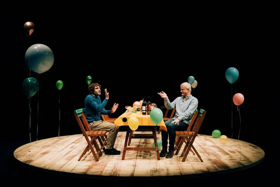 Dos actores sentados a una mesa con globos