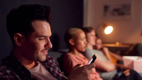 Un joven mirando el m&oacute;vil mientras ve la televisi&oacute;n